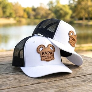 May include: Two white trucker hats with black mesh backs are stacked on a wooden surface. Each hat features a brown leather patch with the word "PAPA" and the names "KRISTIN & LIAM EST. 2026". The hats are near a lake.