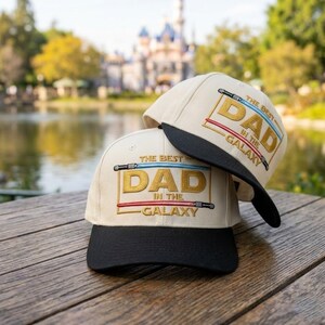 May include: Two beige baseball caps with black brims are stacked on a wooden surface. The caps feature the text "THE BEST DAD IN THE GALAXY" in gold, blue, and red, with lightsaber graphics. The background shows a lake and a castle.