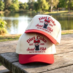 May include: Two off-white baseball caps with red brims, each featuring a red graphic of a glass bottle with a bow, the text Diet Coke, EST. 1885, Social Club, and the slogan Just for the taste of it printed in red on the front panels.