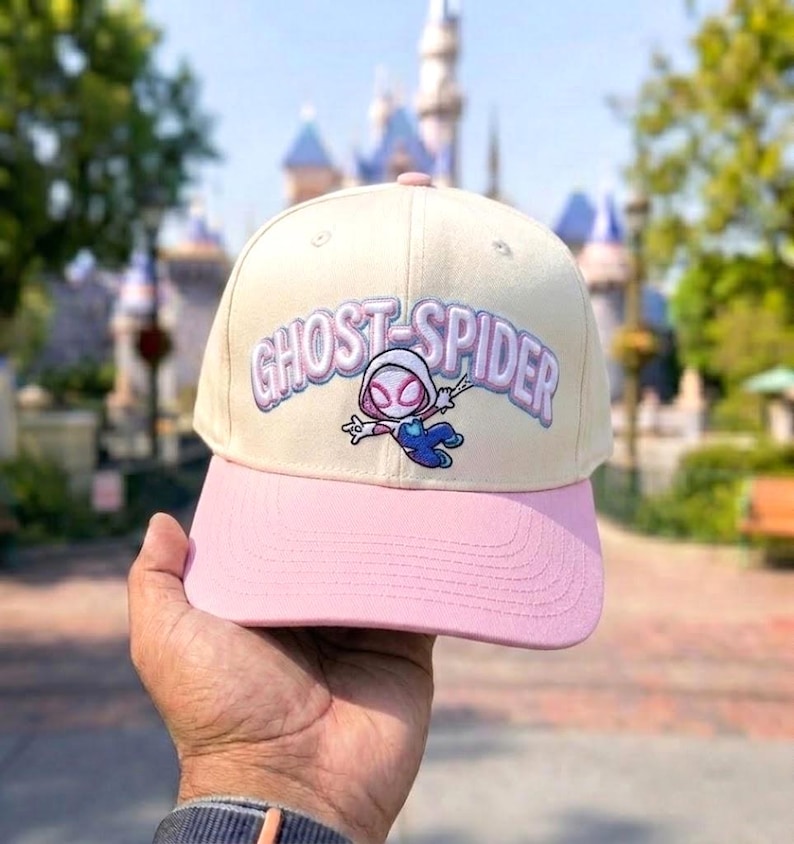 May include: A cream-colored baseball cap with a pink brim featuring an embroidered Ghost-Spider character and the text GHOST-SPIDER in purple and white lettering across the front panel. The cap has a structured crown and stitched eyelets.