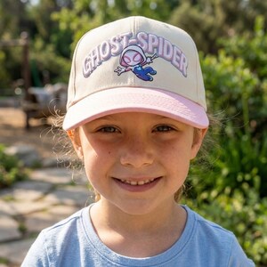 May include: A cream-colored baseball cap with a light pink brim. The front features an embroidered graphic of the character Ghost-Spider in a white, pink, and blue suit, with the text GHOST-SPIDER arched above in purple and white lettering.