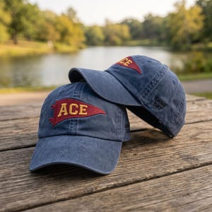 Op de afbeelding: Twee blauwe baseballpetten met een verwassen afwerking. Elke pet heeft een rode wimpelvormige patch met de gele tekst ACE op de voorkant geborduurd. Aan de zijkant van elke pet staat de tekst GAME DAY in zwarte draad op het zijpaneel geborduurd.