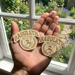 May include: Two Basgiath War College embroidered patches. Each patch features the text "BASGIATH" in a curved banner above a circular emblem with a golden griffin and the text "WAR COLLEGE". The patches are cream-coloured with gold embroidery.