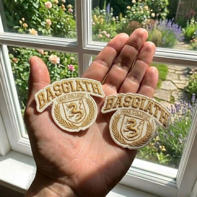 May include: Two Basgiath War College embroidered patches. Each patch features the text "BASGIATH" in a curved banner above a circular emblem with a golden griffin and the text "WAR COLLEGE". The patches are cream-coloured with gold embroidery.