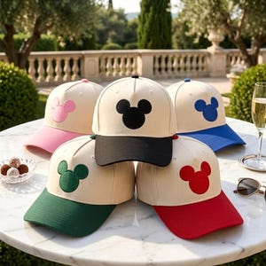 May include: Five baseball caps with cream-coloured crowns and colourful brims. Each cap features a prominent embroidered Mickey Mouse head silhouette on the front panel. The brims and matching icons are pink, black, green, blue, and red respectively.