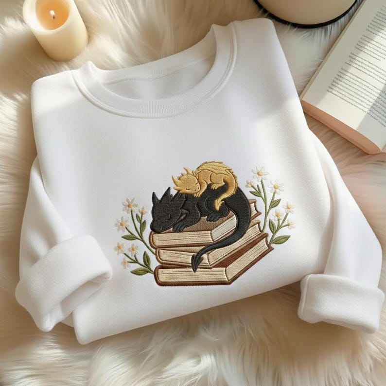 May include: A white crewneck sweatshirt featuring an embroidered design of a black dragon and a smaller gold dragon sleeping on a stack of three books, surrounded by small white flowers with green stems and leaves on both sides of the books.