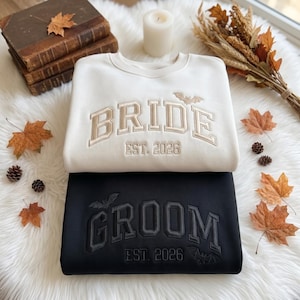 May include: A white sweatshirt with beige embroidered text reading BRIDE EST. 2026 and a small bat icon, stacked on a black sweatshirt with dark gray embroidered text reading GROOM EST. 2026 and two small bat icons, both featuring a collegiate font.