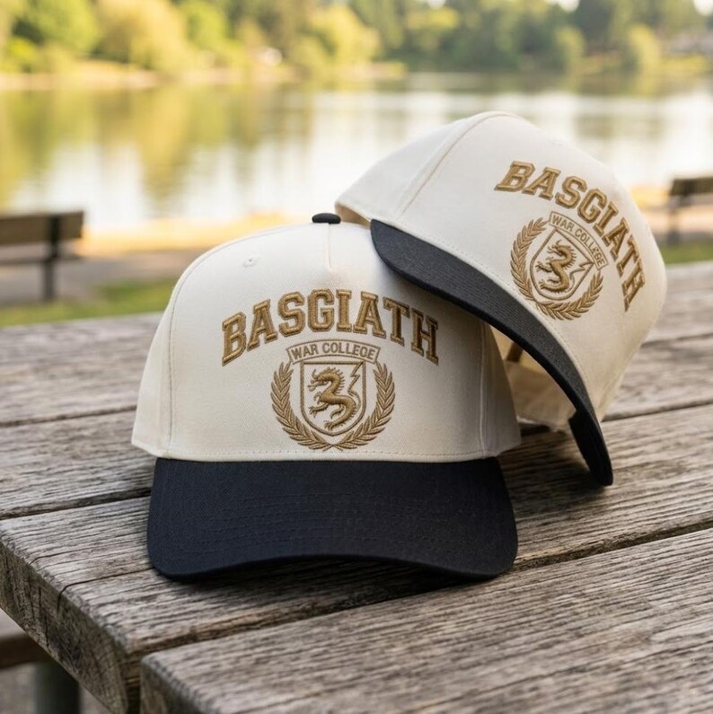 May include: Two baseball caps with the text "BASGIATH WAR COLLEGE" embroidered in gold thread. The caps have a cream-coloured crown and a black brim. The caps are resting on a wooden surface.