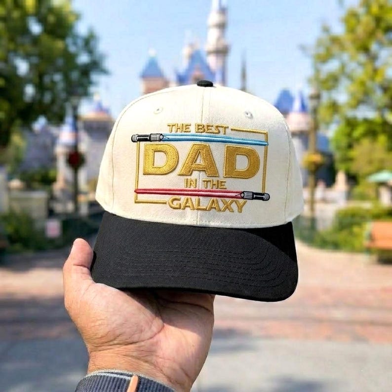 May include: Cream-colored baseball cap with a black brim. The cap features the text "THE BEST DAD IN THE GALAXY" in gold and red, with lightsaber designs. The cap is held up in front of a blurred background.
