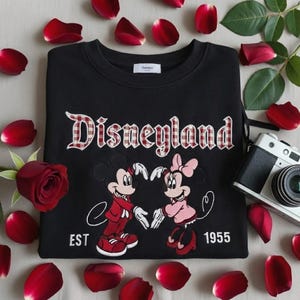 May include: Black sweatshirt with "Disneyland" in a plaid font, featuring Mickey and Minnie Mouse forming a heart. The sweatshirt has "EST 1955" below the characters. Red rose petals and a camera surround the sweatshirt.