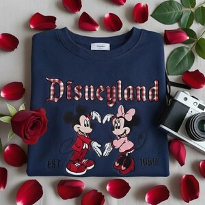 May include: Navy blue Disneyland sweatshirt featuring Mickey and Minnie Mouse holding hands in a heart shape. The word "Disneyland" is embroidered in red and white checkered letters. The sweatshirt has the year "EST 1955" embroidered below the characters.