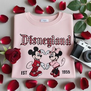 May include: A light pink Disneyland sweatshirt featuring Mickey and Minnie Mouse forming a heart with their hands. The word "Disneyland" is in a plaid font. The sweatshirt has the text "EST 1955". Red rose petals and a camera surround the sweatshirt.