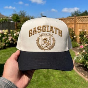 May include: A baseball cap with a cream-coloured crown and a black brim. The cap features the embroidered text "BASGIATH WAR COLLEGE" in gold, along with a crest of a dragon and laurel leaves. The cap is held up against a background of green foliage and a blue sky.