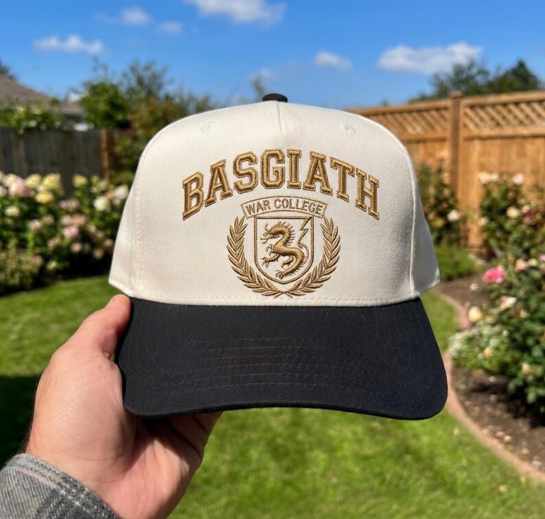May include: A baseball cap with a cream-coloured crown and a black brim. The cap features the embroidered text "BASGIATH WAR COLLEGE" in gold, along with a crest of a dragon and laurel leaves. The cap is held up against a background of green foliage and a blue sky.