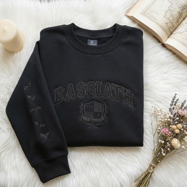 May include: Black crew neck sweatshirt featuring the word "BASGIATH" in arched lettering. Below is a crest with "WAR COLLEGE". The left sleeve has embroidered dragon silhouettes. The sweatshirt is on a white, fluffy surface, with a candle and open book visible.