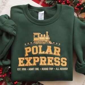 May include: A dark green sweatshirt with the words "POLAR EXPRESS" in gold lettering, along with a gold train graphic. The text "EST. 1998 - ADMIT ONE - ROUND TRIP - ALL ABOARD!" is printed below the main text. The sweatshirt has a crew neck and long sleeves.