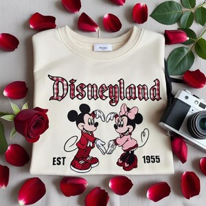 May include: Cream-colored sweatshirt featuring the word "Disneyland" in a red plaid font, with Mickey and Minnie Mouse illustrations. The design includes the text "EST 1955". The sweatshirt is surrounded by red rose petals and a vintage camera.
