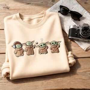 May include: Cream-colored sweatshirt featuring a cartoon graphic of four Grogu characters in various poses. One Grogu wears headphones, another holds a ball, and a third has a question mark above its head. The sweatshirt is on a wooden surface.