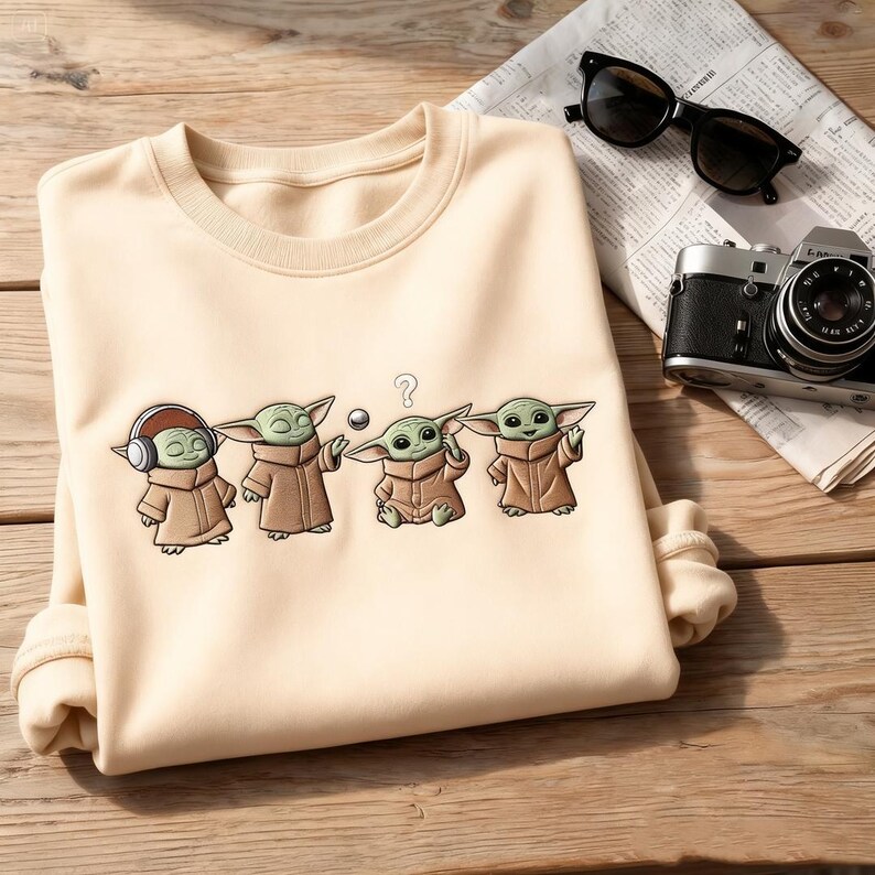 May include: Cream-colored sweatshirt featuring a cartoon graphic of four Grogu characters in various poses. One Grogu wears headphones, another holds a ball, and a third has a question mark above its head. The sweatshirt is on a wooden surface.