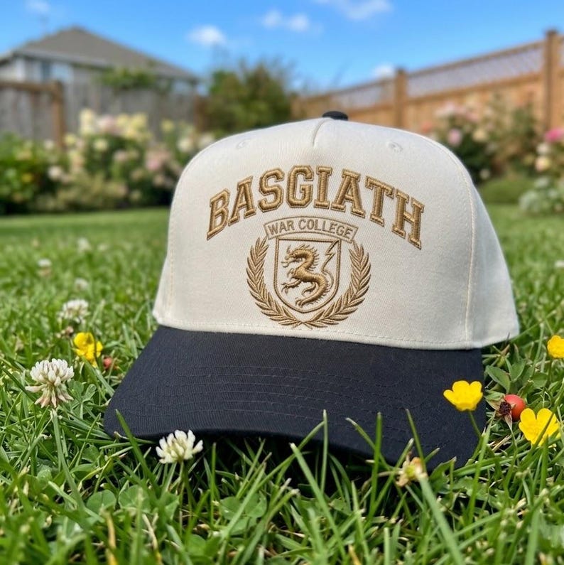 May include: A cream and navy blue baseball cap with gold embroidery. The cap displays the text "BASGIATH WAR COLLEGE" and a dragon emblem. The hat is set on green grass, with a blurred background of a garden and a blue sky.