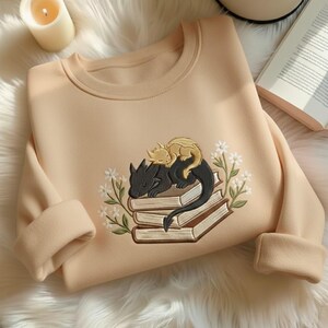 May include: A beige crewneck sweatshirt featuring an embroidered design of a black dragon and a smaller yellow dragon sleeping on a stack of three books, surrounded by white flowers with green stems and leaves on both sides of the illustration.