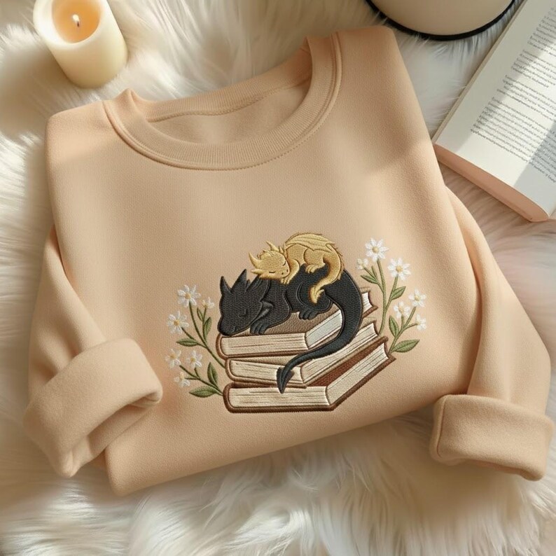May include: A beige crewneck sweatshirt featuring an embroidered design of a black dragon and a smaller yellow dragon sleeping on a stack of three books, surrounded by white flowers with green stems and leaves on both sides of the illustration.