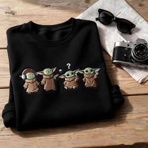 May include: Black crewneck sweatshirt featuring an embroidered design of four Grogu characters in various poses. The design includes a Grogu with headphones, one with a ball, one with a question mark, and one with a peace sign. A vintage camera and sunglasses are also visible.