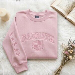 May include: A pale pink sweatshirt with the word "BASGIATH" embroidered across the chest in a collegiate style. The left sleeve features embroidered dragon silhouettes. A shield emblem is below the text. The sweatshirt is on a white fluffy surface.