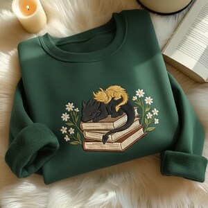 May include: A dark forest green crewneck sweatshirt featuring an embroidered design of a small gold dragon resting on a larger black dragon, both curled atop a stack of three books, surrounded by white daisy flowers with green stems and leaves.