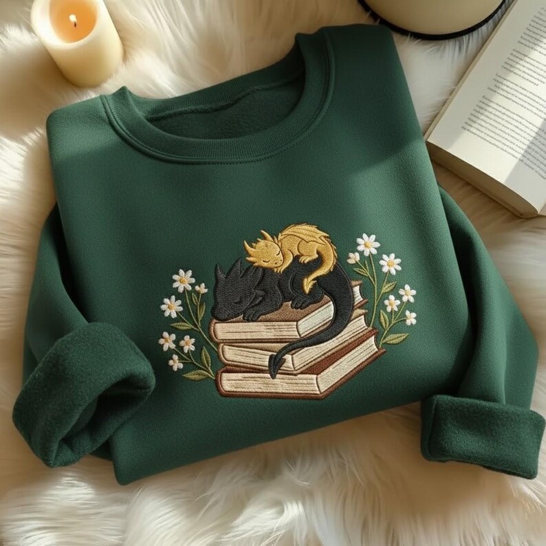 May include: A dark forest green crewneck sweatshirt featuring an embroidered design of a small gold dragon resting on a larger black dragon, both curled atop a stack of three books, surrounded by white daisy flowers with green stems and leaves.