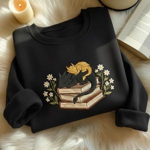 May include: A black crewneck sweatshirt featuring an embroidered design of a small gold dragon resting on top of a larger black dragon, both curled up on a stack of three books surrounded by white flowers with green stems and leaves.