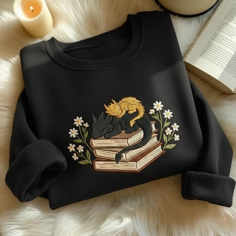 May include: A black crewneck sweatshirt featuring an embroidered design of a small gold dragon resting on top of a larger black dragon, both curled up on a stack of three books surrounded by white flowers with green stems and leaves.