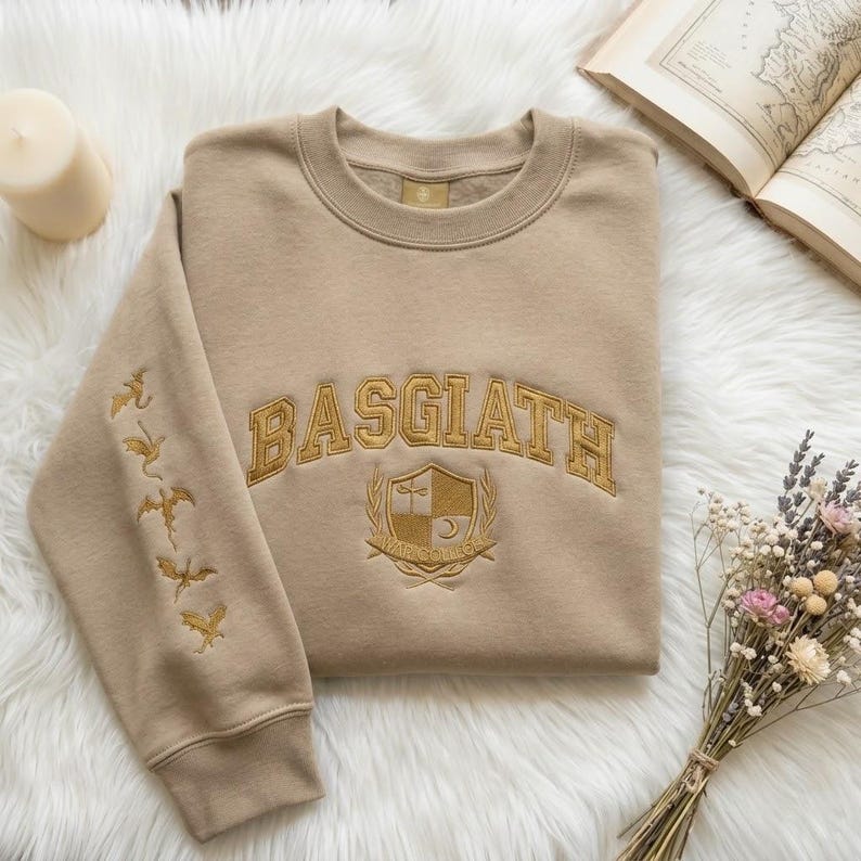 May include: Beige crew neck sweatshirt with gold embroidered text "BASGIATH" and a crest design. The left sleeve has gold dragon embroidery. A candle, open book, and flowers are in the background.