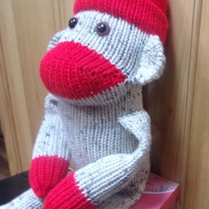 Pode incluir: Um brinquedo de macaco de meia de malha com um chapéu vermelho e braços e pernas vermelhos. O macaco está sentado em uma caixa rosa com desenhos florais.