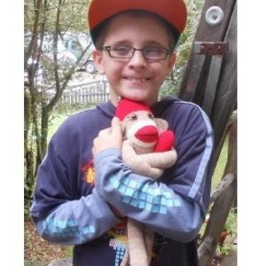 May include: A young person wearing a red and orange baseball cap and a blue long-sleeved shirt is holding a sock monkey. The monkey is brown with red accents and has a white face.