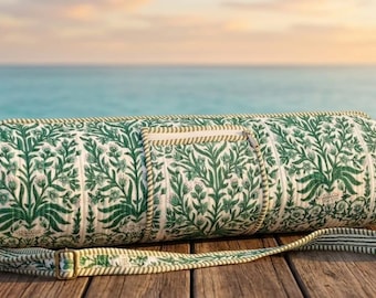 May include: A green and white floral patterned yoga mat bag. The cylindrical bag has a matching shoulder strap and is made of quilted fabric. It rests on a wooden surface, with a beach scene in the background.