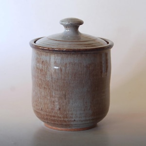 May include: A cylindrical ceramic canister with a matching lid, featuring a mottled glaze that blends earthy brown tones with soft grey hues. The surface has a smooth, glossy finish with subtle horizontal ridges near the top rim.