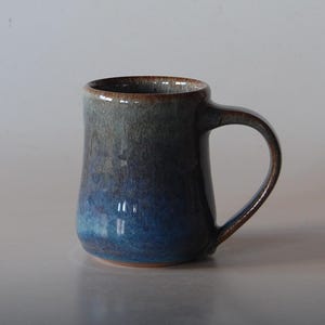 May include: A handmade ceramic mug with a gradient blue glaze. The mug has a rounded body, a sturdy handle, and a brown rim. This stoneware mug is perfect for coffee, tea, or any hot beverage.