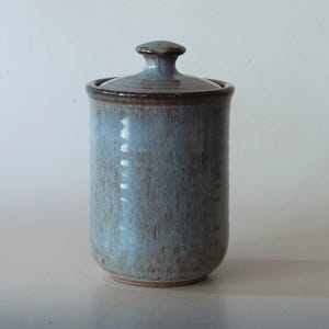 May include: A cylindrical ceramic canister with a matching lid, featuring a mottled light blue and brown glaze. The surface has a textured appearance with vertical streaks of color, and the lid is topped with a small, rounded knob handle.