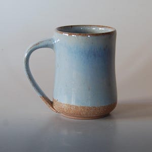 May include: A handmade ceramic mug with a light blue glaze, transitioning to a darker blue towards the top. The base and handle are a natural, unglazed brown. The mug has a curved handle and a cylindrical shape.