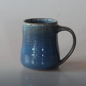 May include: A handmade ceramic mug with a blue ombre glaze. The mug has a rounded base, a cylindrical body, and a large, curved handle. The glaze transitions from a lighter blue at the rim to a darker blue at the base. The mug is suitable for hot or cold beverages.
