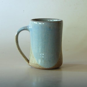 May include: A cylindrical ceramic mug featuring a two-tone glaze. The upper portion is finished in a smooth, light blue glaze, while the lower section and the curved handle reveal a textured, speckled tan clay body with a natural, earthy appearance.