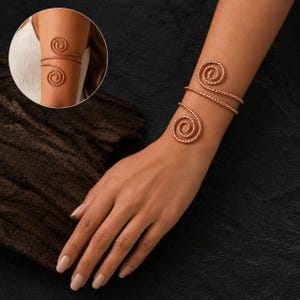 May include: A rose gold-toned arm cuff featuring a twisted metal wire design that wraps around the arm. The piece consists of two circular spiral ends connected by a single diagonal strand, creating an open, adjustable silhouette for the upper arm.