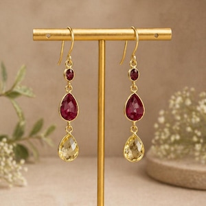 May include: Gold-tone earrings with a three-stone design. Each earring features a small, round red gemstone at the top, a large, pear-shaped red gemstone in the middle, and a pear-shaped yellow gemstone at the bottom. The earrings are displayed on a gold stand.