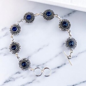 May include: Silver-toned bracelet featuring seven circular, dark blue gemstone settings. Each setting is surrounded by a detailed, decorative border. The bracelet has a toggle clasp closure and is displayed on a white marble surface.