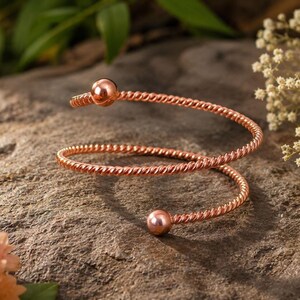 May include: A rose gold-toned spiral bracelet featuring a twisted metal wire design. The bracelet has a circular, open-ended shape with a smooth, polished spherical bead at each of the two ends, creating a continuous, coiled appearance.