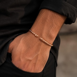 Könnte beinhalten: Ein roségoldfarbenes Armband in Manschettenform mit einer gedrehten, seilartigen Textur. Das Armband ist offen gestaltet und verfügt über abgerundete, glatte Enden, die ein durchgehendes Spiralmuster entlang des Metallbandes bilden.