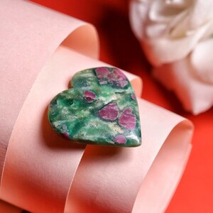 May include: A heart-shaped gemstone, predominantly green with red inclusions. The polished stone has a natural, organic look. It rests against a backdrop of pink ribbon and a red surface. The gemstone is a decorative item.