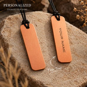 May include: Two rectangular copper-toned pendants with rounded corners, each suspended from a black cord. One pendant is blank, while the other features the engraved text "YOUR NAME". The image includes the text "PERSONALIZED ENGRAVE YOUR NAME".
