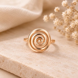 Könnte beinhalten: Ein Ring aus poliertem Roségold mit einer markanten kreisförmigen Vorderseite, die ein durchgehendes, erhabenes Spiraldesign aufweist, das sich nach innen zum Zentrum hin windet. Die Ringschiene ist glatt und mit der Rückseite verbunden.